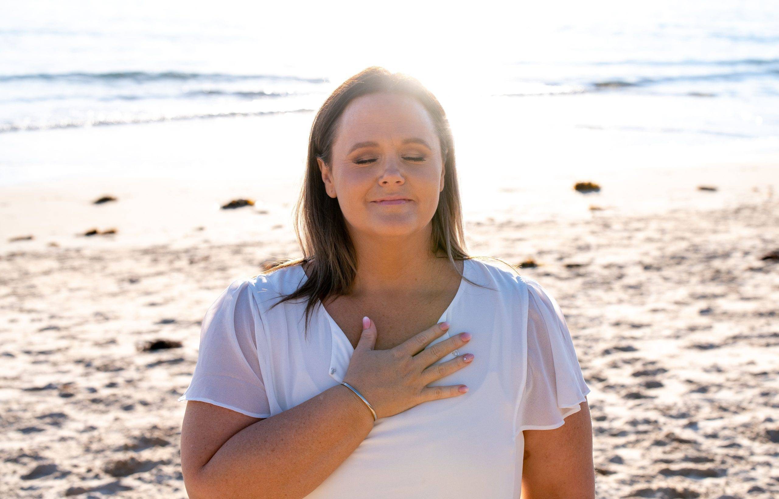 Holly with her hand to her chest on the beach with the sun behind her